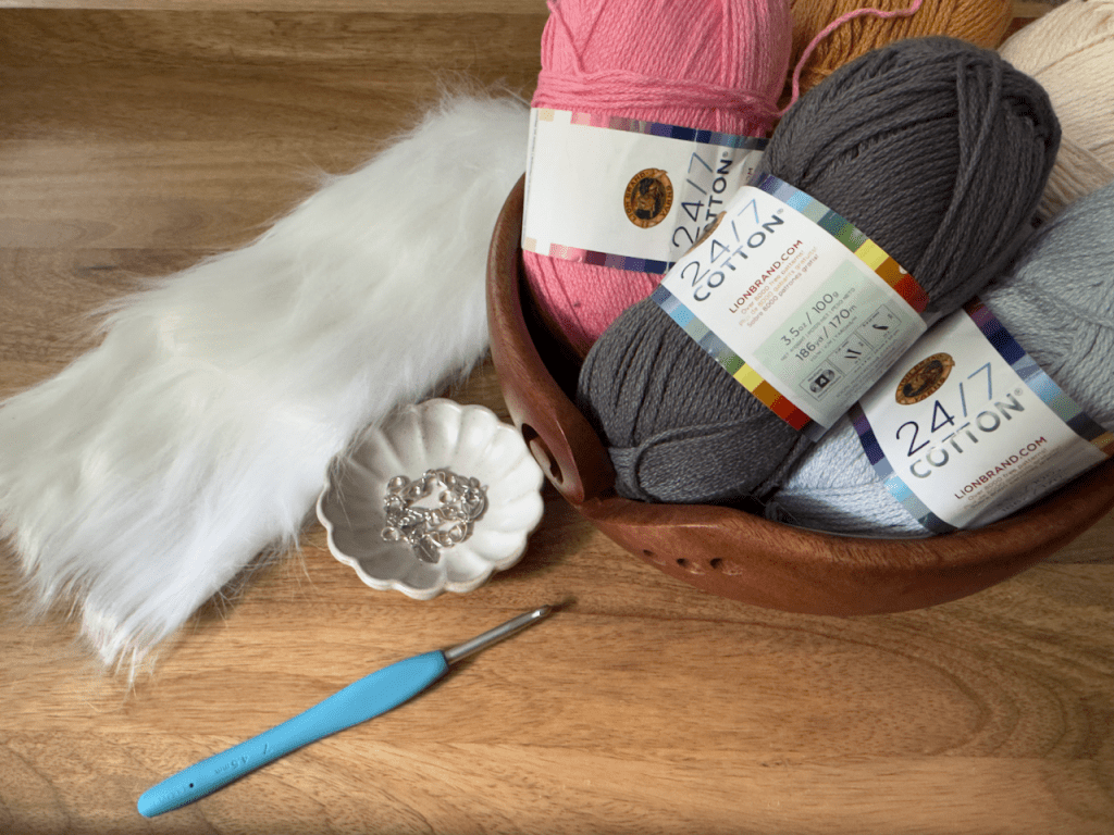 a blue 4.5mm crochet hook, faux fur, stitch markers, and skeins of yarn in pink, dark gray, light gray, and camel yellow