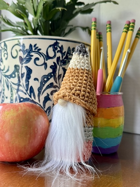 crochet gnome in the shape and color of a pencil with white beard and beige felted wool nose. there is a potted plant, a red apple, and a cup of pencils and crochet hooks in the background.